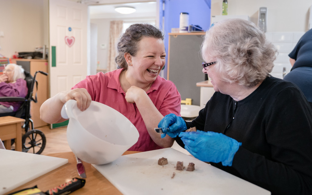 Two individuals with gloves doing an activity at a table with a person in a wheelchair in the background.