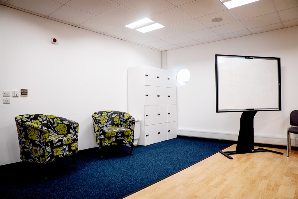 Bright meeting room featuring two floral chairs, storage lockers, a large whiteboard, and carpeted flooring.